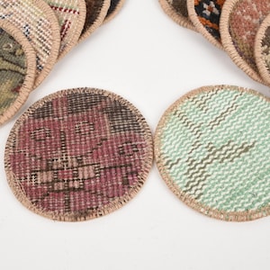 May include: Circular coasters with a woven rug design. One coaster features a red and black pattern, while another has a green and white zig-zag pattern. The coasters have a beige border and are arranged on a white surface.