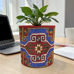 May include: A cylindrical plant pot with a vibrant, patterned design. The pot features a blue background with a red and cream-colored geometric pattern. The top rim is red, and the pot holds a green leafy plant.