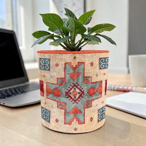 Puede incluir: Una maceta cilíndrica beige con un patrón geométrico rojo y turquesa, que contiene una planta verde. La maceta tiene un borde rojo y está sobre un escritorio de madera. Un portátil y una taza están en el fondo.
