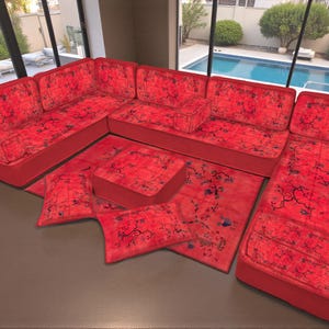 May include: A sectional sofa set in a vibrant red hue, featuring a floral pattern in darker shades. The set includes a corner sofa, a rug, and several cushions. The furniture is arranged in a modern living space with large windows.