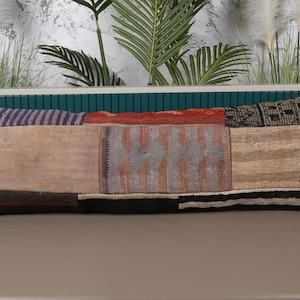 May include: Rectangular decorative pillow with a patchwork design. The pillow features various fabric sections in shades of beige, red, and blue. The pillow is approximately 30 inches long and 12 inches wide, with a black and white striped border.