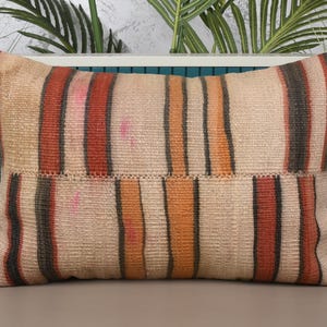 May include: Rectangular beige pillow with vertical stripes in shades of red, orange, and black. The pillow has a textured appearance, suggesting a woven fabric. The design is simple and rustic, suitable for home decor.