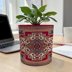 May include: A cylindrical plant pot with a detailed, patterned fabric design. The fabric features a central medallion in blue, cream, and red, reminiscent of a traditional rug. The pot holds a green leafy plant, adding a touch of nature to the design.
