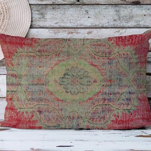 Puede incluir: Cojín rectangular con un diseño descolorido de estilo vintage. El cojín presenta un motivo floral central en tonos verdes y beige, rodeado por un borde rojo. La estética general recuerda a una alfombra desgastada.