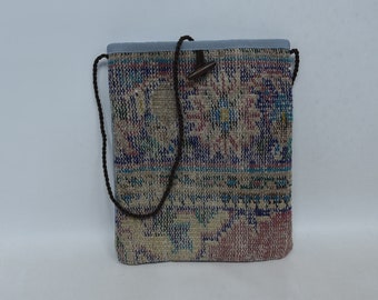 May include: A rectangular tote bag with a woven, patterned design in shades of blue, pink, and beige. The bag has a light blue top and a brown cord strap. A wooden toggle closure is visible.