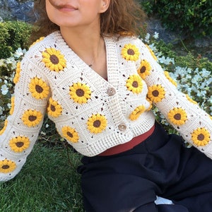 May include: A cream-colored crocheted cardigan with a sunflower design. The cardigan has a V-neck and is cropped. The model is wearing the cardigan with a black skirt.