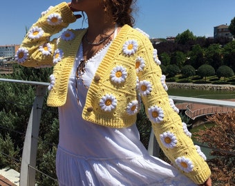 May include: A yellow crocheted cardigan with white daisy flowers. The cardigan is cropped and has long sleeves.