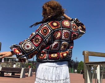 May include: A colorful crochet cardigan with a geometric pattern in shades of red, orange, yellow, white, and black. The cardigan is made of a soft, textured yarn and has a relaxed fit.