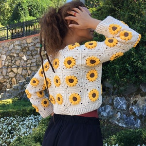 May include: A white crocheted cardigan with a sunflower pattern. The cardigan has long sleeves and a cropped length.