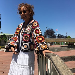 May include: A colorful crocheted cardigan with a black background and a geometric pattern of squares. The cardigan has long sleeves and a loose fit.