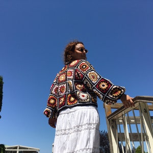 May include: A woman wearing a colorful crocheted cardigan with a white skirt. The cardigan is made of squares with different floral patterns in red, yellow, and white.