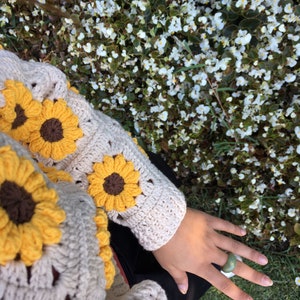 May include: A crocheted sweater with a sunflower design in shades of yellow and brown. The sweater is worn by a person with a green ring on their finger.