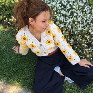 May include: A white crocheted cardigan with yellow sunflower accents. The cardigan is cropped and has a button closure. The model is wearing the cardigan with a black skirt.