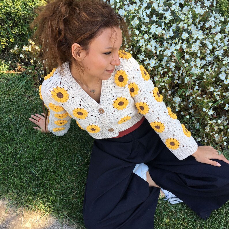 May include: A white crocheted cardigan with yellow sunflower accents. The cardigan is cropped and has a button closure. The model is wearing the cardigan with a black skirt.