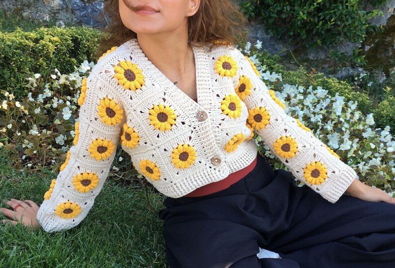 May include: A cream-colored crocheted cardigan with a sunflower design. The cardigan has a V-neck and is cropped. The model is wearing the cardigan with a black skirt.