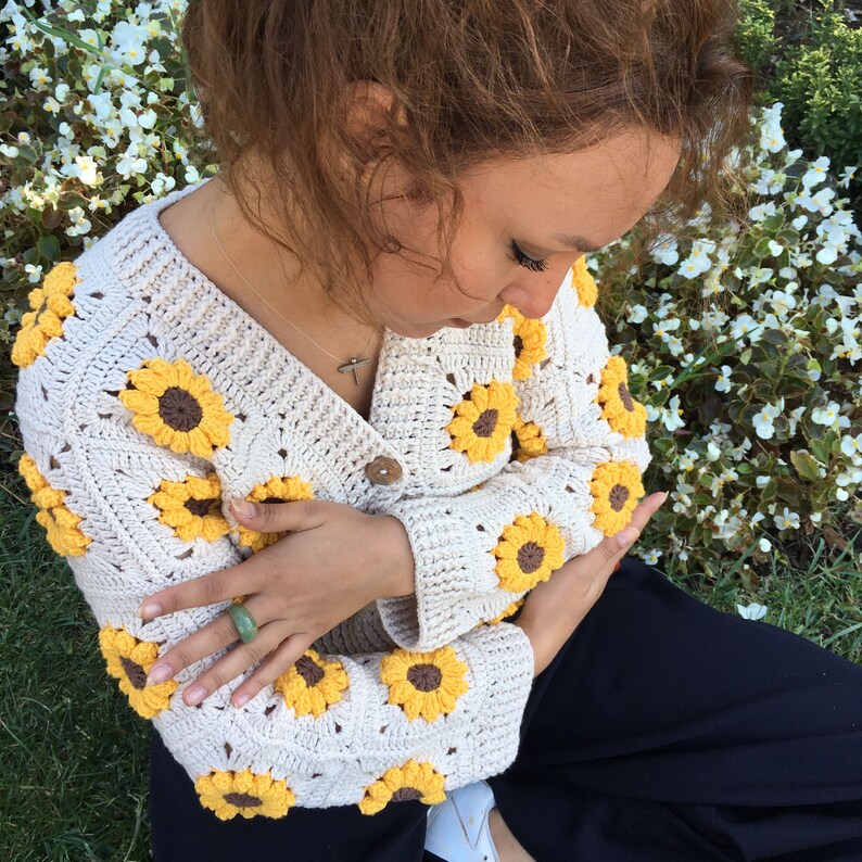 May include: A cream-colored crocheted cardigan with a sunflower design. The cardigan has a button closure and is made with a soft, textured yarn.