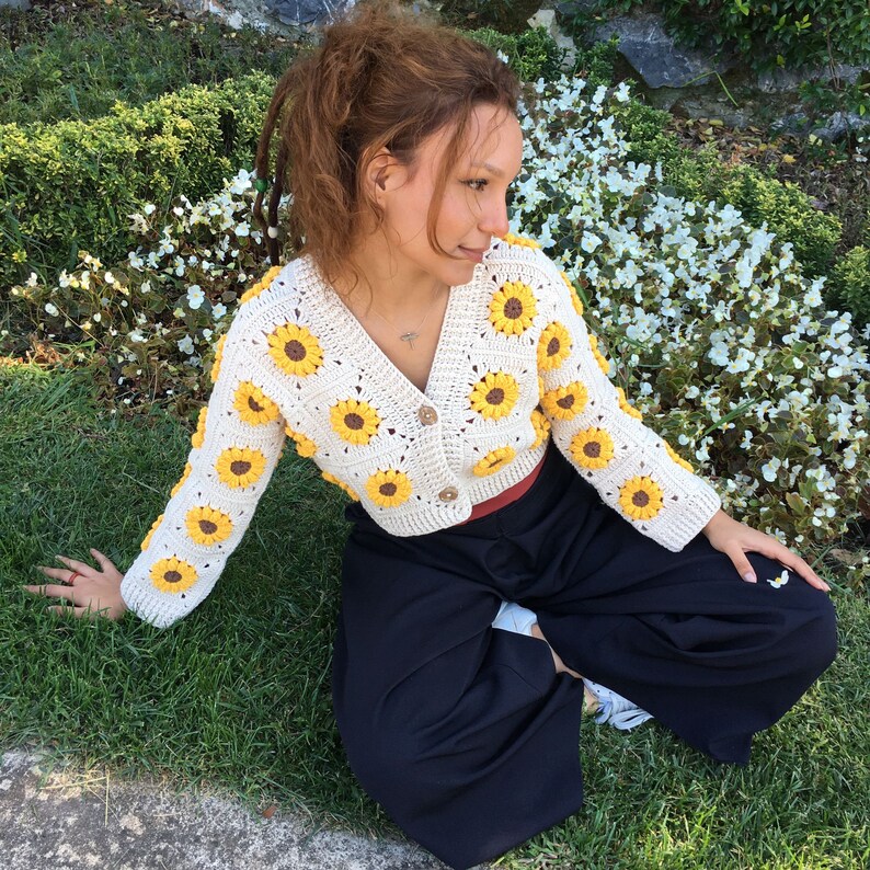 May include: A white crocheted cardigan with yellow sunflower accents. The cardigan is cropped and has a button closure. The model is wearing the cardigan with black pants.