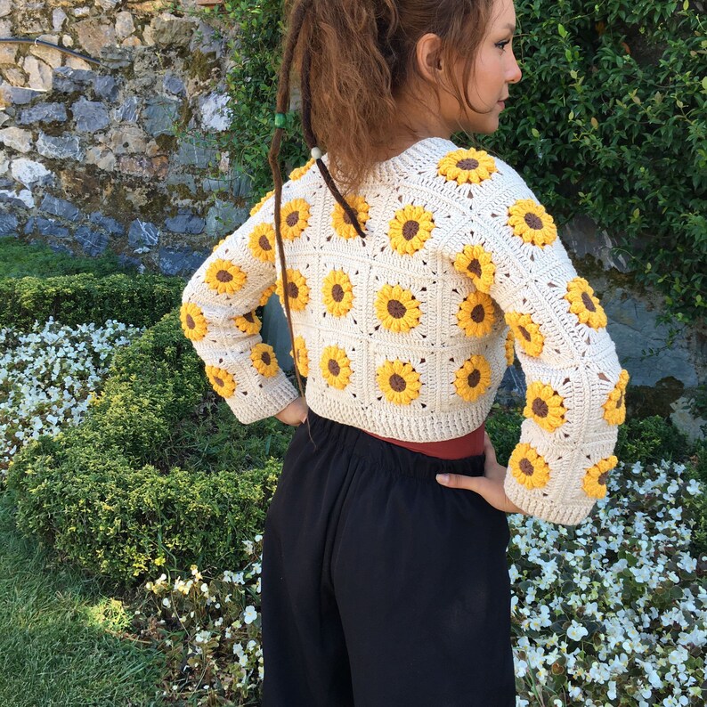 May include: A white crocheted cropped cardigan with a sunflower pattern. The cardigan has long sleeves and a round neckline.