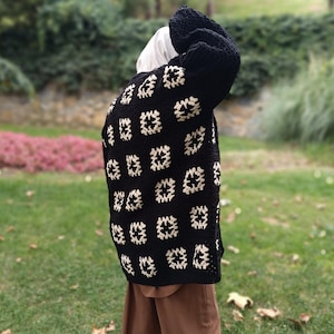 May include: A black crochet cardigan with a repeating square pattern in cream. The cardigan has long sleeves and a relaxed fit. The person is wearing brown pants and a white head covering.