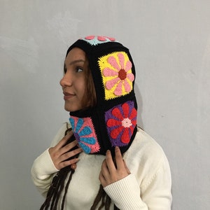 May include: A black crochet hooded hat with colorful flower designs. The hat has a long neck piece and is made with a chunky yarn.
