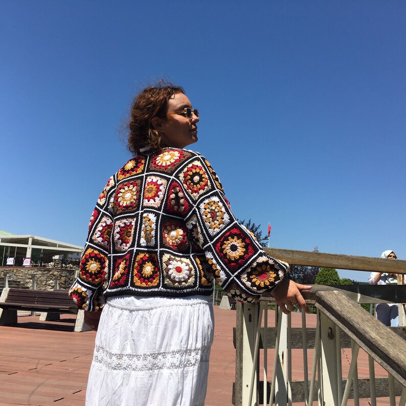 May include: A woman wearing a colorful crocheted cardigan with a white skirt. The cardigan is made of squares with different colors and patterns. The woman is standing on a wooden deck with a railing in the background.