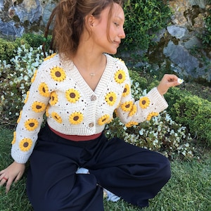 May include: A white crocheted cardigan with a sunflower pattern. The cardigan has a V-neck and button closure. The model is wearing black pants.