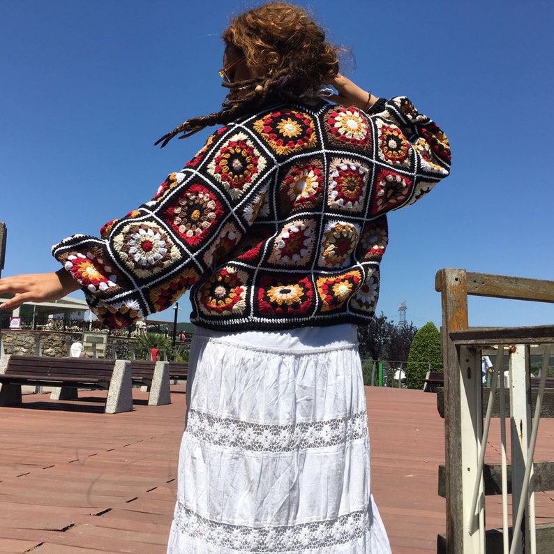 May include: A colorful crochet cardigan with a geometric pattern in shades of red, orange, yellow, white, and black. The cardigan is made of a soft, textured yarn and has a relaxed fit.