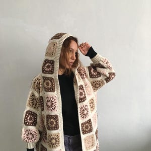 May include: A hooded, open-front crochet cardigan in a patchwork design. The sweater features square panels in cream, brown, and pink, with a floral pattern in the center of each square. The sleeves are long and the hood is also made of the same crochet squares.
