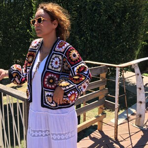 May include: A woman wearing a white dress and a crocheted cardigan with a colorful geometric pattern. The cardigan has a black background with red, yellow, white, and brown squares.