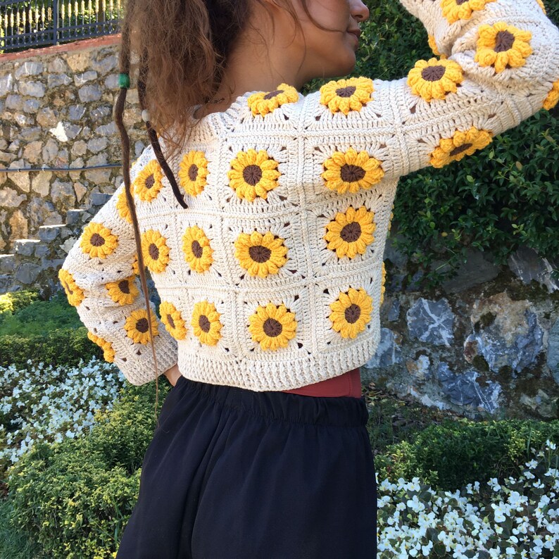 May include: A white crocheted cardigan with sunflower motifs. The cardigan is cropped and has long sleeves.