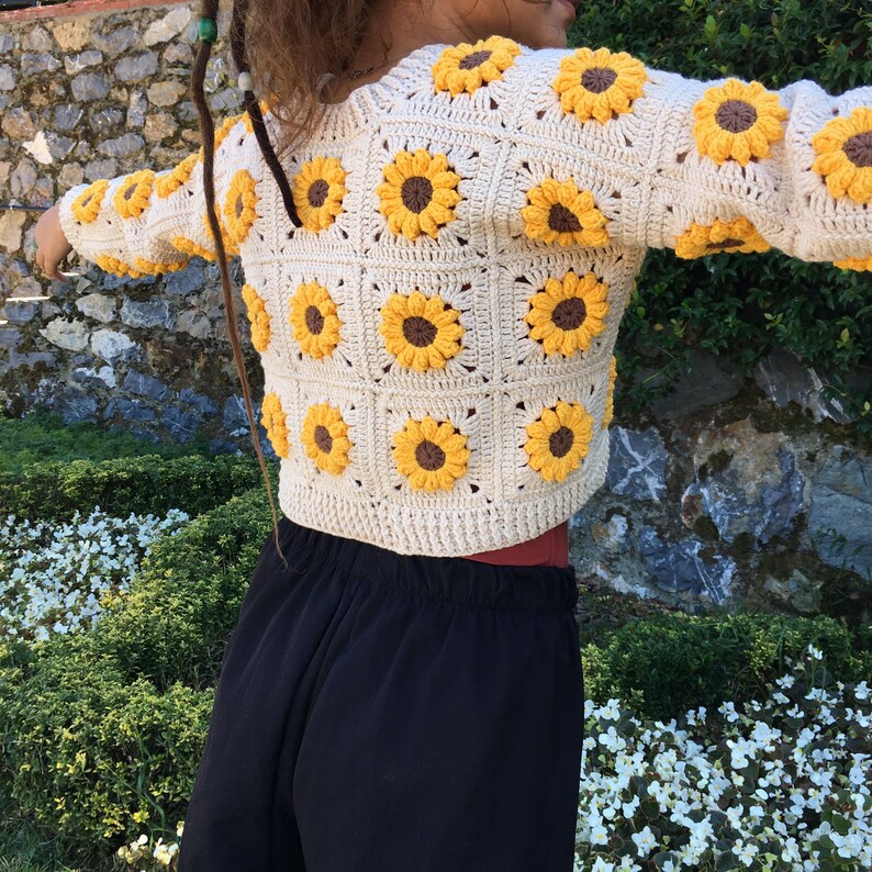 May include: A cream-colored crocheted cardigan with a sunflower pattern. The cardigan is cropped and has long sleeves.