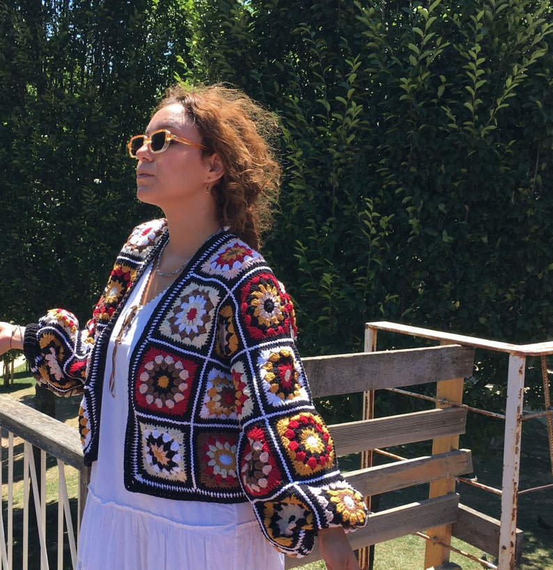 May include: A colorful crocheted jacket with a black background and a variety of colored squares. The jacket has a V-neck and long sleeves.
