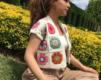 May include: A white crocheted vest with colorful floral granny squares. The vest has short sleeves and a V-neck.