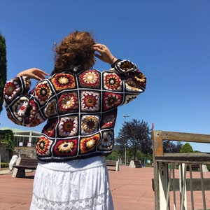 May include: A colorful crochet cardigan with a square pattern in shades of red, brown, white, and yellow. The cardigan has a loose fit and long sleeves.