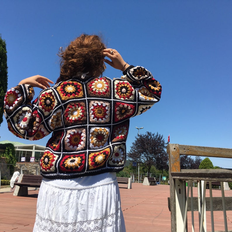 May include: A colorful crochet cardigan with a square pattern in shades of red, brown, white, and yellow. The cardigan has a loose fit and long sleeves.