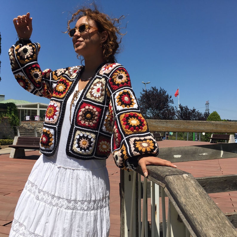 May include: A colorful crocheted jacket with a geometric pattern in shades of red, orange, yellow, white, and black. The jacket is open in the front and has long sleeves.