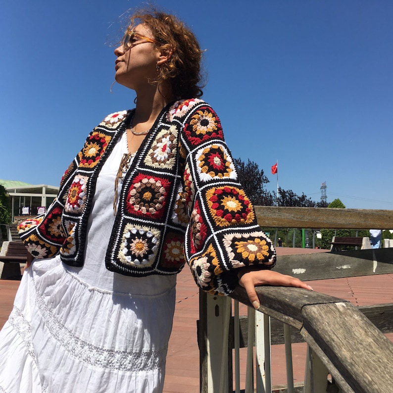 May include: A colorful crocheted cardigan with a geometric pattern in shades of red, orange, yellow, white, and black. The cardigan has a loose fit and long sleeves.