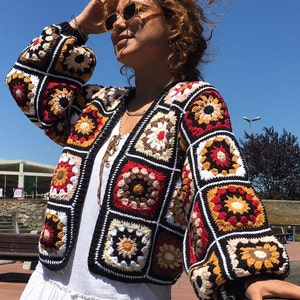 May include: A colorful crocheted cardigan with a geometric pattern in shades of red, orange, yellow, brown, white, and black. The cardigan has a loose fit and a long sleeve.