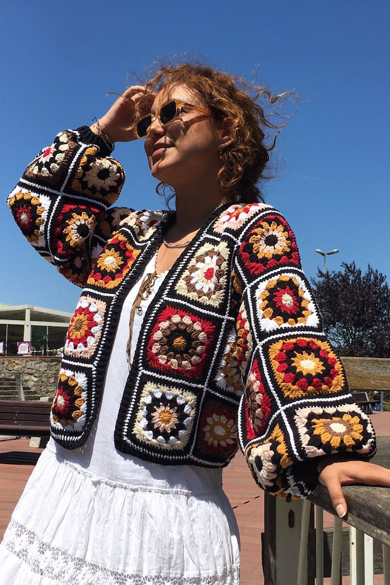 May include: A colorful crocheted cardigan with a geometric pattern in shades of red, orange, yellow, brown, white, and black. The cardigan has a loose fit and a long sleeve.