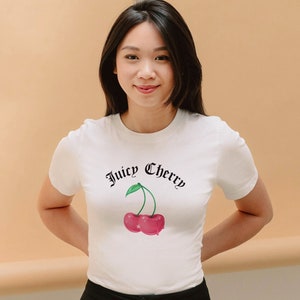 May include: White t-shirt featuring the text "Juicy Cherry" in a gothic font above a graphic of a red cherry with a green leaf. The shirt is short-sleeved and worn by a person with dark hair.