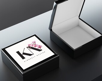 May include: Black square box with a white lid. The lid has a floral design with the letters "KN" and the text "KIMBERLY NGUYEN".