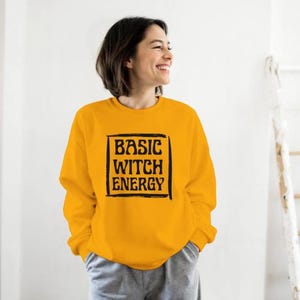May include: Mustard yellow sweatshirt with the text "BASIC WITCH ENERGY" in a black, bold font. The text is enclosed in a black rectangular frame. The sweatshirt is long-sleeved and has a crew neck.