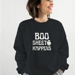 May include: Black sweatshirt with the text "BOO SHEET HAPPENS" in white, along with a ghost graphic. The sweatshirt is a crew neck style and is being worn by a person.