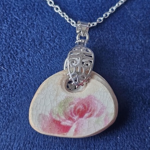 May include: A silver chain necklace with a pendant featuring a delicate floral design on a white ceramic base. The pendant is shaped like a heart and has a pink rose design.