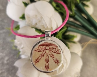 May include: A white ceramic pendant with a red and white design of a bow and arrows. The pendant is on a pink cord.