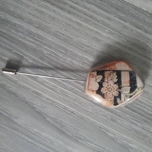 Puede incluir: Un alfiler de plata con una cabeza de cerámica hexagonal de color crema con un diseño floral en blanco y negro.