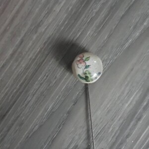 May include: A white porcelain pin with a floral design in pink, green, and blue. The pin is attached to a silver metal pin back.