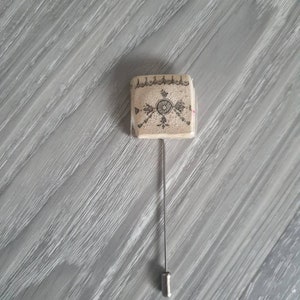 May include: A square-shaped pin with a beige background and a black and white geometric design. The pin has a silver metal stem.