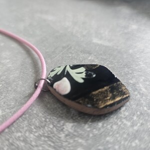 May include: A black and gold pendant necklace with a floral design. The pendant is shaped like a teardrop and has a pink flower in the centre. The necklace is made of a pink cord.