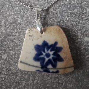 Pode incluir: Um pingente de cerâmica branca com um desenho floral azul, pendurado em uma corrente de prata.